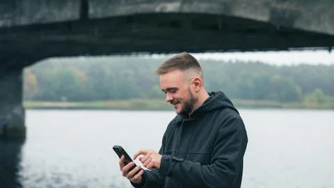 A Man Smiles While Using a Smartphone Under a Bridge by the River During a Stock Photos