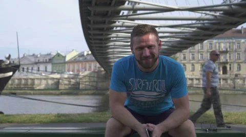 Man is smiling to camera sitting on the bench against the river. Vidéo 65188600