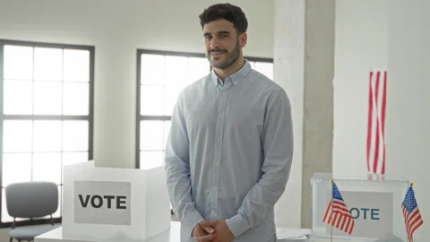 Man smiling with hands clasped by a ballot box and vote sign, standing at a.. Stock Footage 339175551