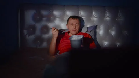 Man is smiling lying on the bed in front of the TV with a bucket of ice cream Stock Footage 132827533