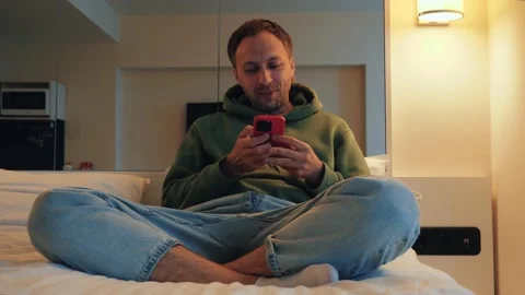 Man smiling with phone in hands lying on bed at home. Young guy making video Video stock 239253853