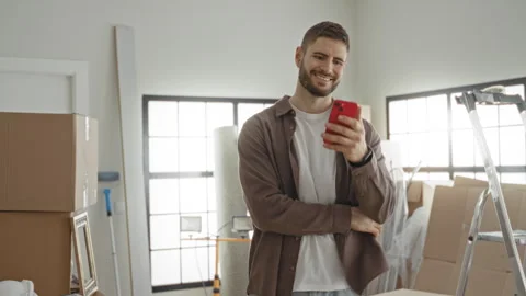 Man smiling while holding red smartphone amid packed moving boxes in buildi.. Stock Footage 337366555