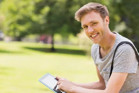 Man smiling while tapping tablet on park grass wearing grey t-shirt and backpack Stock Photos