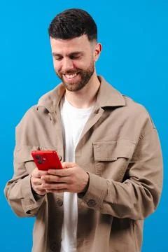 Man smiling while using a red smartphone against a bright blue background Foto stock