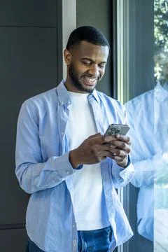 Man smiling while using smartphone by a bright window, casually dressed in .. Fotos Stock