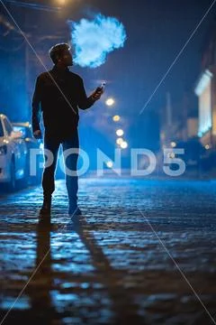 The man smoke electrical cigarette on the street. Evening night time Stock Photos