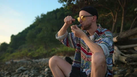 Man smokes marijuana from pipe and blows fume sitting on shore Stock Footage 233608115