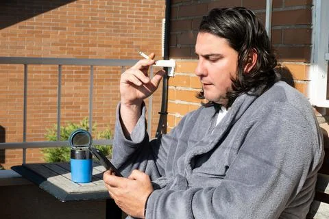 Man smoking cigarette on balcony using smartphone in bathrobe Foto stock