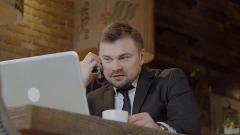 Man solves business issues in cafe on the phone sitting in front of laptop Vídeos de archivo 70592901