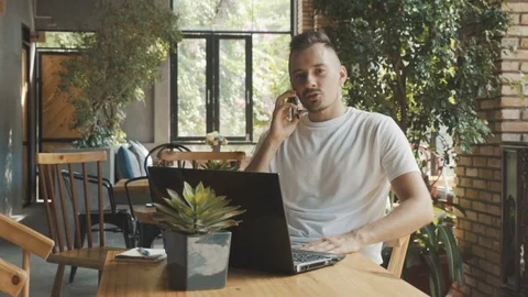 Man speaks with friend on smartphone while working on computer in cafe. Stock Footage 76491644