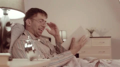 Man in spectacles is sitting in bed with a book and laughing his pleasure 库存影片 68420867
