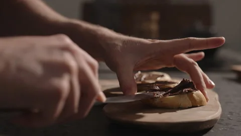 Man spreading chocolate hazelnut cream on baguette slice on olive board Stock Footage 148411922