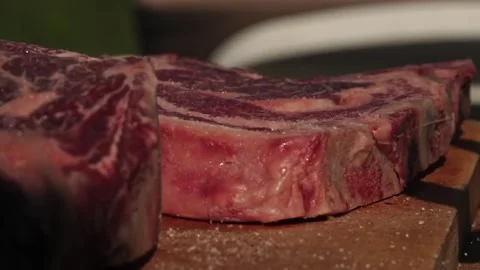 Man sprinkling salt over a raw rib eye steak on a wooden board Video stock 250157998