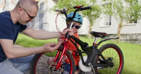 Man squats down pointing at bicycle and explaining task to son 動画素材 285497502