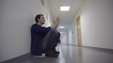 Man squats in the long white corridor, leaning against the wall, praying, hoping Stock Footage 72432670
