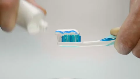A man squeezes toothpaste from a tube onto a blue toothbrush on a white back Stock Footage 130423423