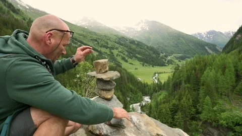 A man stacking rocks to build a cairn overlooking a scenic mountain valley with Stockbeeldmateriaal 281045470