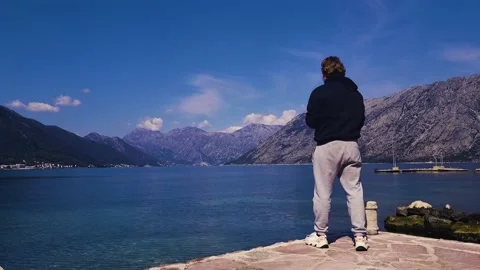 A man standing and throwing stones into the water with a scenic mountain view Stock Footage 237998753