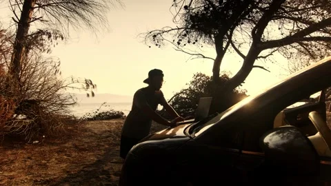 Man Standing and Working on Laptop on Car Hood by the Sea at Sunset Stockbeeldmateriaal 341939581