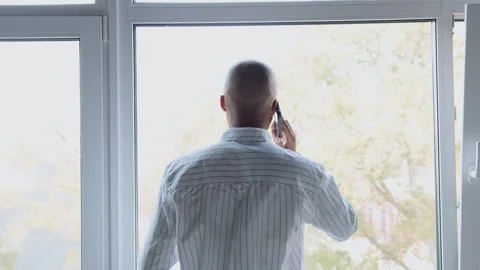 Man standing with back to camera, looking through window while talking on phone, Stock Footage 335755536