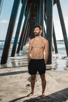 Man standing barefoot on beach looking at the ocean under a pier Photos