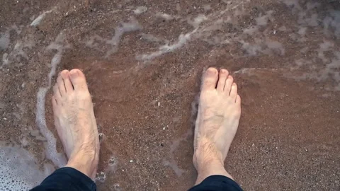 Man standing barefoot on the sand with waves washing over his feet 스톡 동영상 347774528