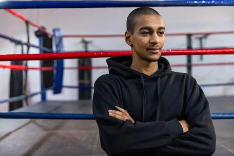 Man standing in boxing ring in black hoodie, arms crossed, observing heavy bag, Stock Photos