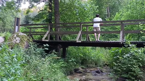 A man is standing on a bridge on a summer day, looking at the mountain river Stock Footage 262928190