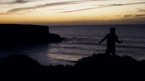 Man standing on cliff and throwing stone in sea during sunset Stock Footage 71542158