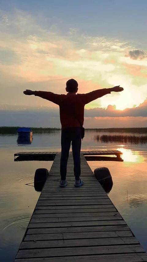 A man standing on a dock next to a body of water, refracted sunset Stock Footage 280892403
