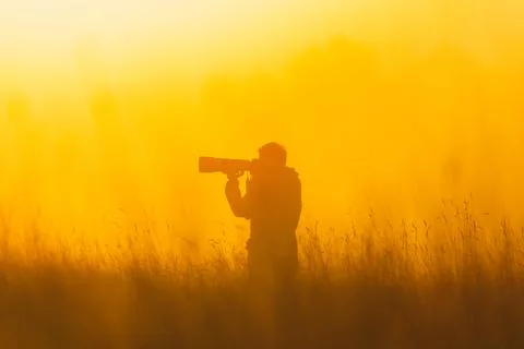 A man standing in a field with a camera, capturing the beauty of the sunset. Stock Photos