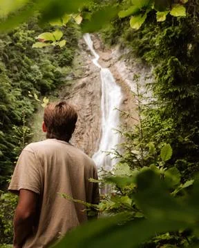 Man Standing in Forest Looking at Tall Cascading Waterfall Stockfoto's