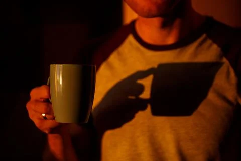 A man standing in front of a window in sunset lighting is drinking coffee in  Stock-Fotos