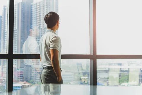 Man standing with hands in pockets looking out of big window at city building. Stock Photos