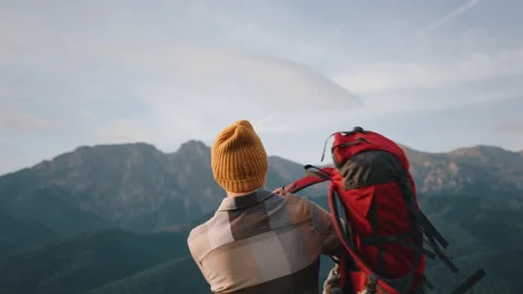 Man standing on hill top overlooking epic mountain peaks, hiking, red backpack Stock Footage 283029629