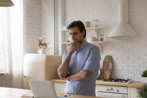Man standing in kitchen staring at laptop thinks over task 스톡 사진