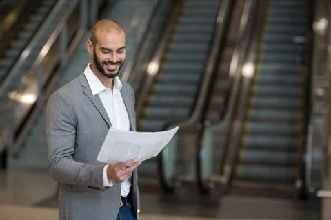 Man standing in large lobby reading folded newspaper near parallel escalators Stockfoto's