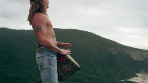 Man standing on a mountain by the ocean and playing the Djembe drums Stock Footage 279026402