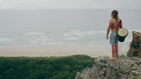 Man standing on a mountain by the ocean and holding djembe drums Stock Footage 279057723