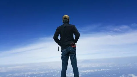 Man standing on a mountain top looking out at the clouds and blue sky Stock Photos