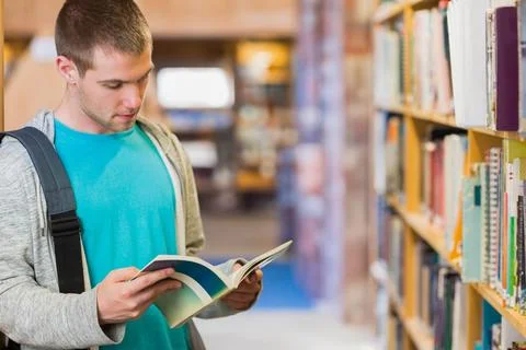 Man standing reading open paperback book in library wearing teal tee, backpack, Stock Photos