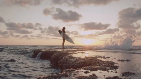 Man standing on a rock platform along the ocean at sunset with his surfboard Stock Footage 275162414