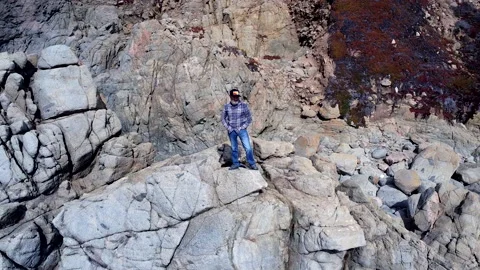 Man Standing on rocks next to ocean as drone flys around him Video stock 217827126