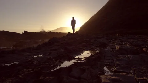 Man standing on rocks by the ocean and waves at sunset Stock Footage 279796603