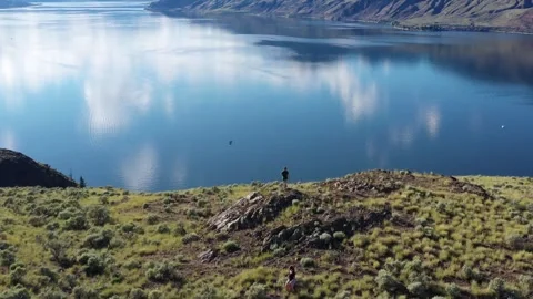 Man Standing on Rocky Cliff Overlooking Reflective Lake Vista in British Stock Footage 334251033