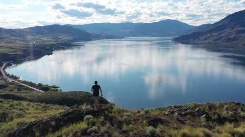 Man Standing on Rocky Cliff Overlooking Reflective Lake Vista in British Stock Footage 334251049