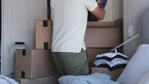 A man is standing in a room with boxes and clothes. New home concept. 動画素材 315701011