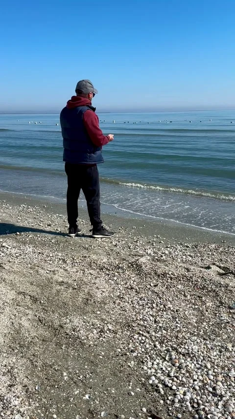 Man standing on a sandy beach feeding seagulls by the sea Stock Footage 336814645