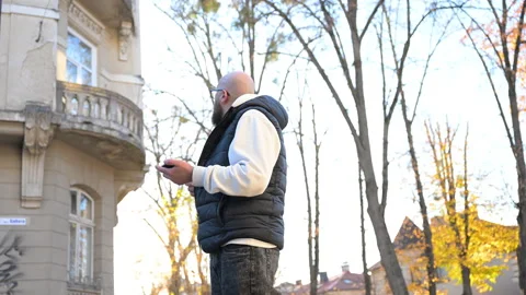 Man standing on sidewalk checking smartphone surrounded by autumn trees and Stock Footage 346546212
