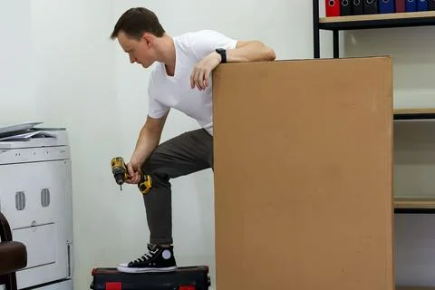Man standing on toolbox drilling into large cardboard box in office Stock Photos